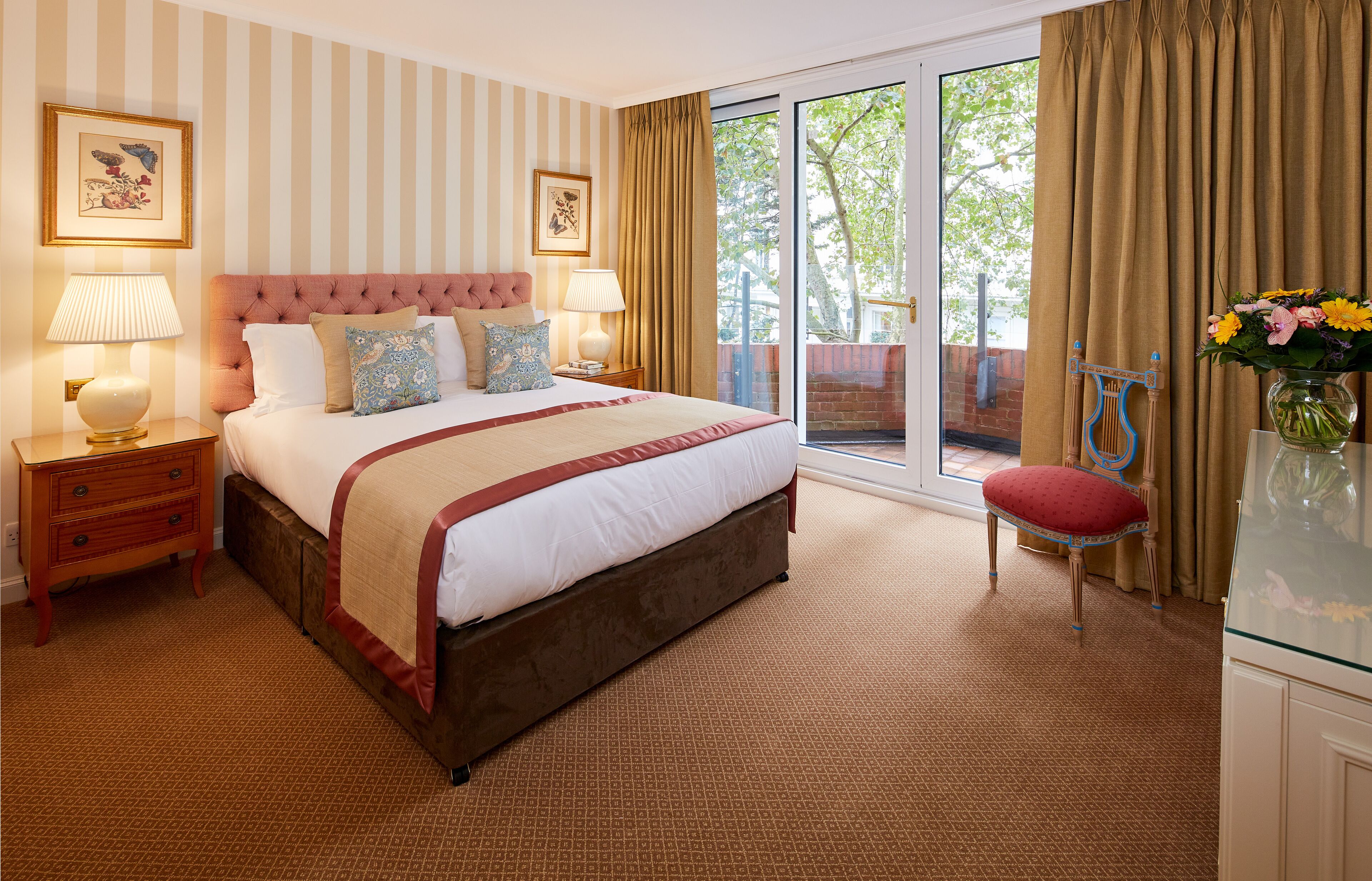 A bedroom with a large bed, two bedside tables, a red chair, a glass cabinet, and a view of the outdoors.