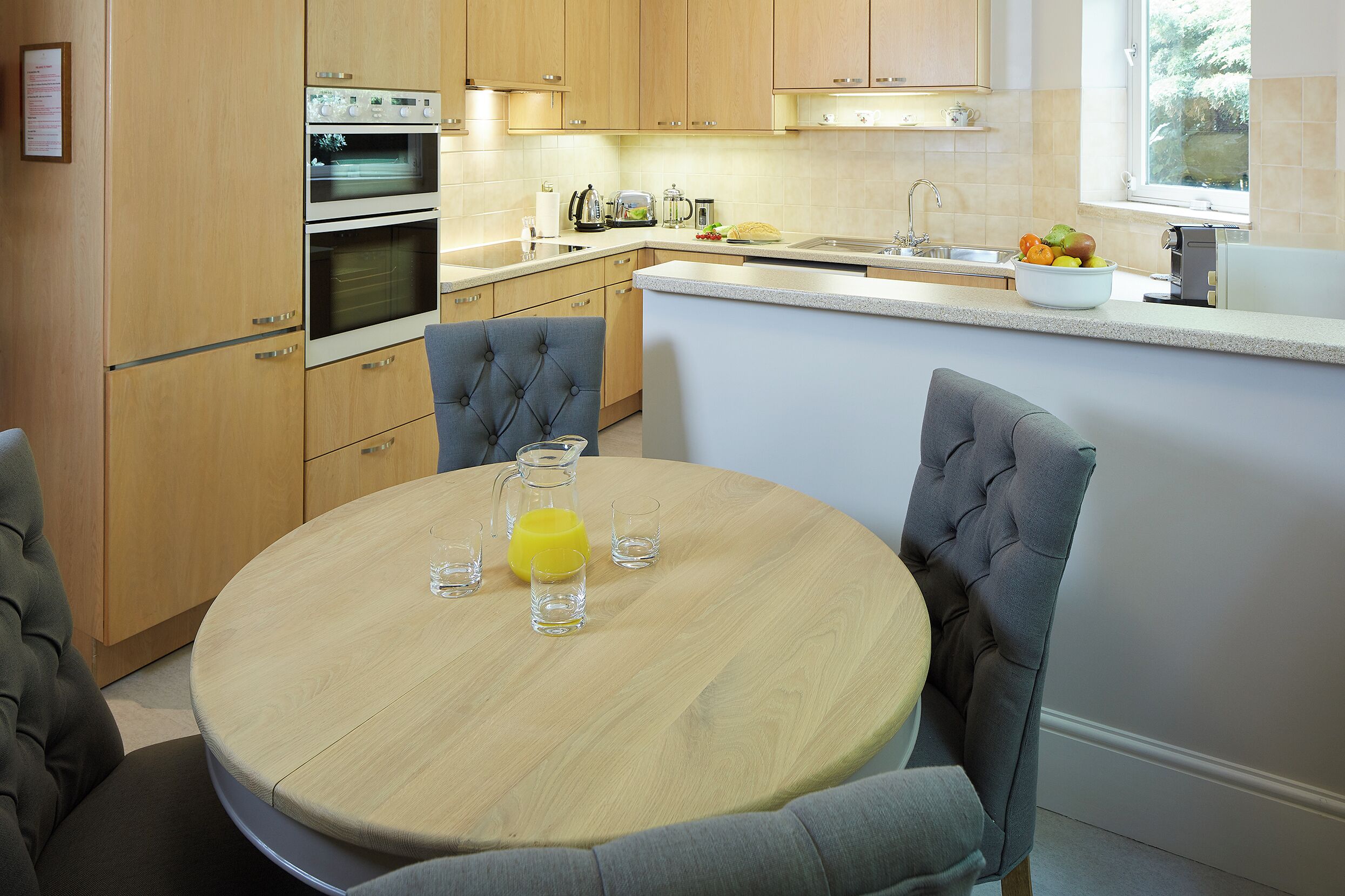 A modern kitchen with wooden cabinets, a double oven, and a round dining table with chairs.