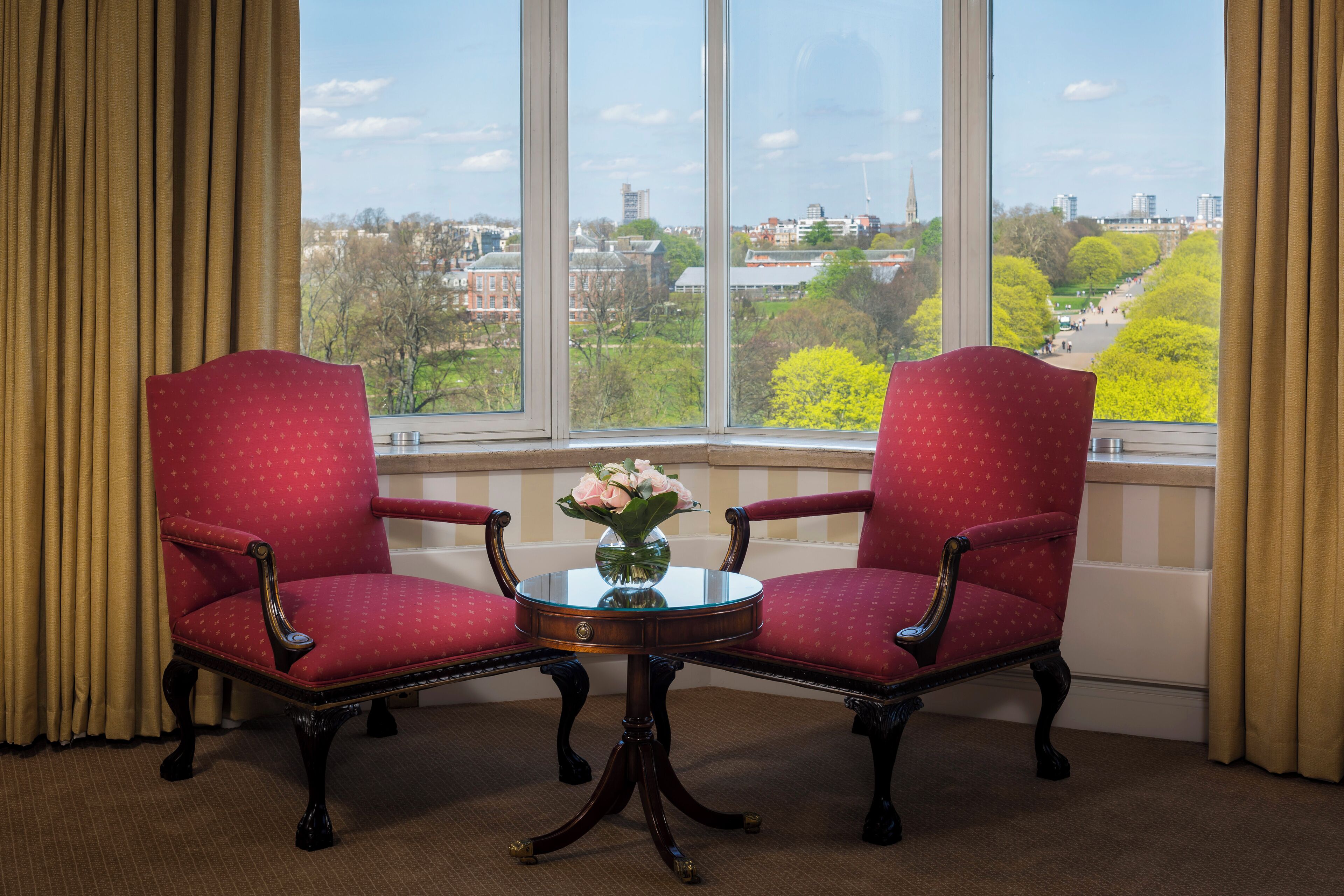 A room with two red upholstered chairs, a small round table with a vase of flowers, and a large window offering a view of a cityscape and park.