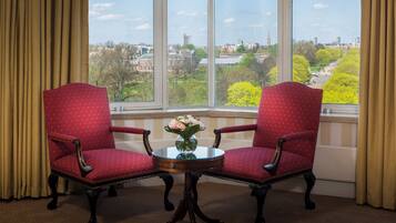 A room with two red upholstered chairs, a small round table with a vase of flowers, and a large window offering a view of a cityscape and park.