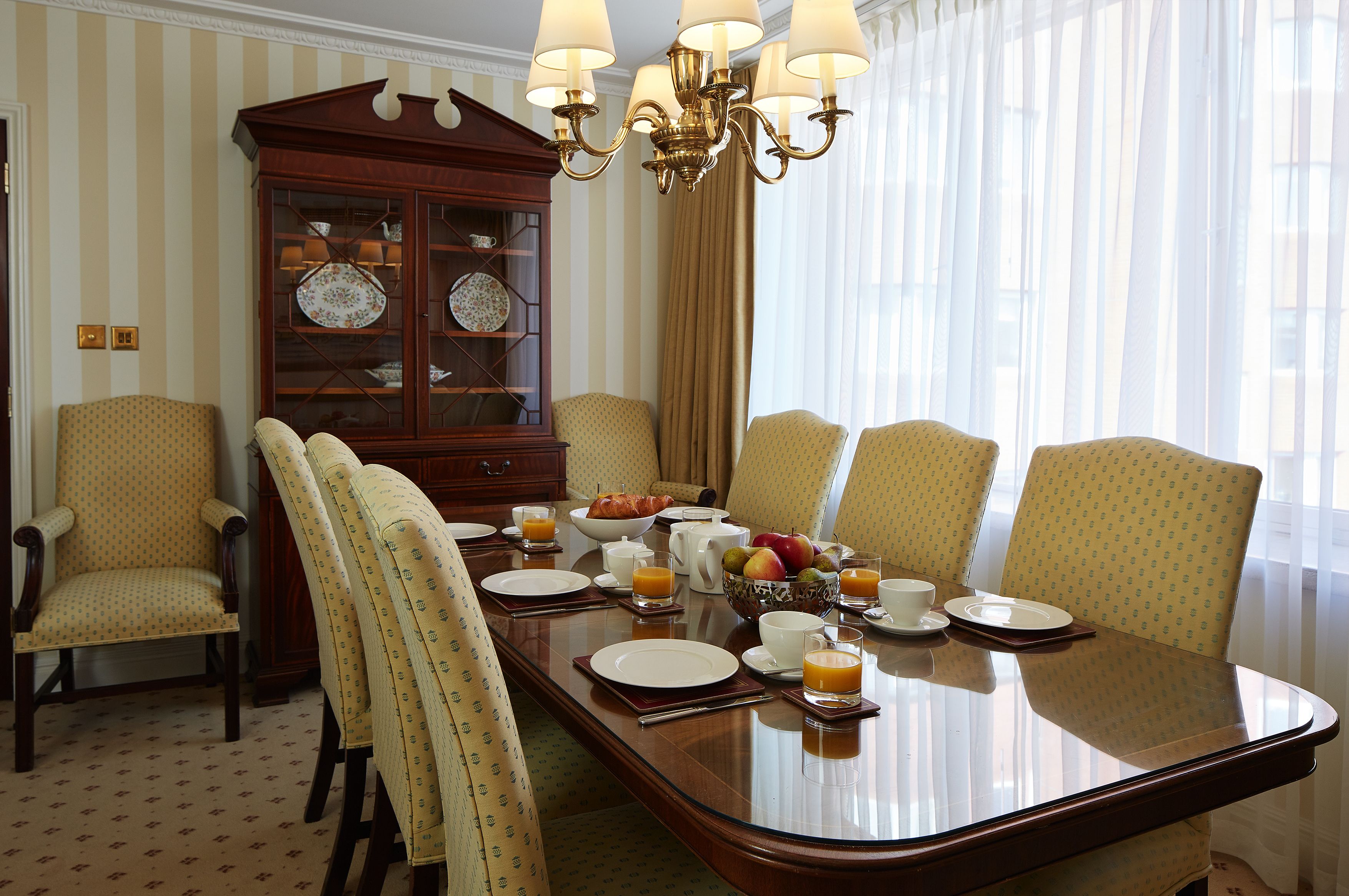 A dining room with a large wooden table set for breakfast, surrounded by patterned chairs, a china cabinet, and a chandelier.