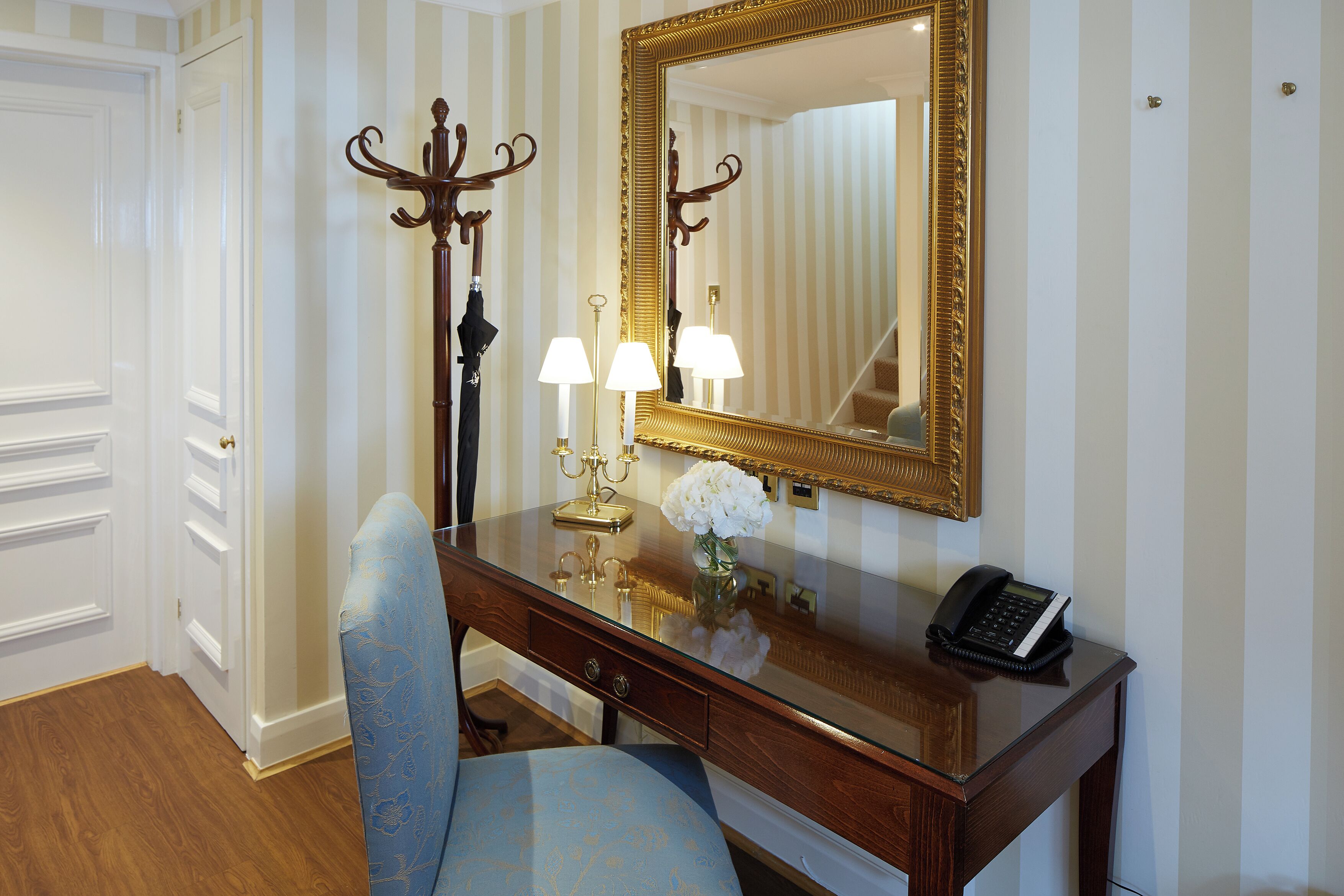 Una habitación de hotel con un escritorio de madera, una silla azul, un espejo grande, dos lámparas y un teléfono.