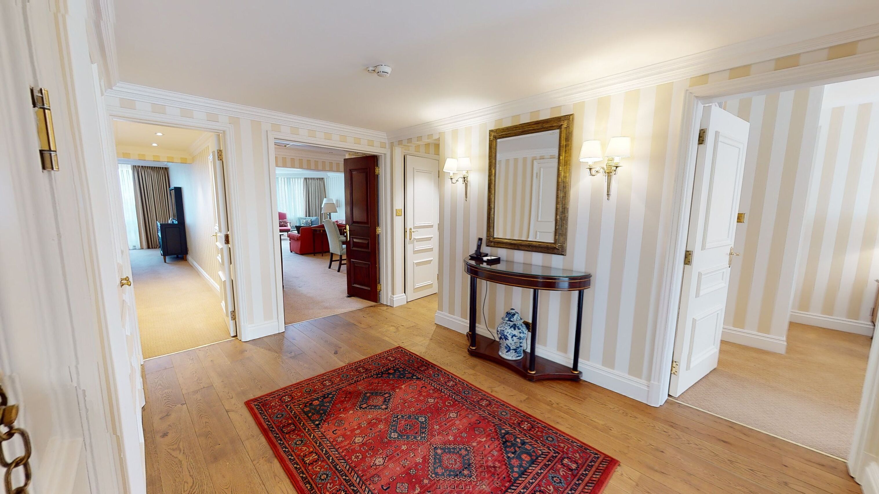 A hallway with a red patterned rug, a console table with a mirror, and striped wallpaper.
