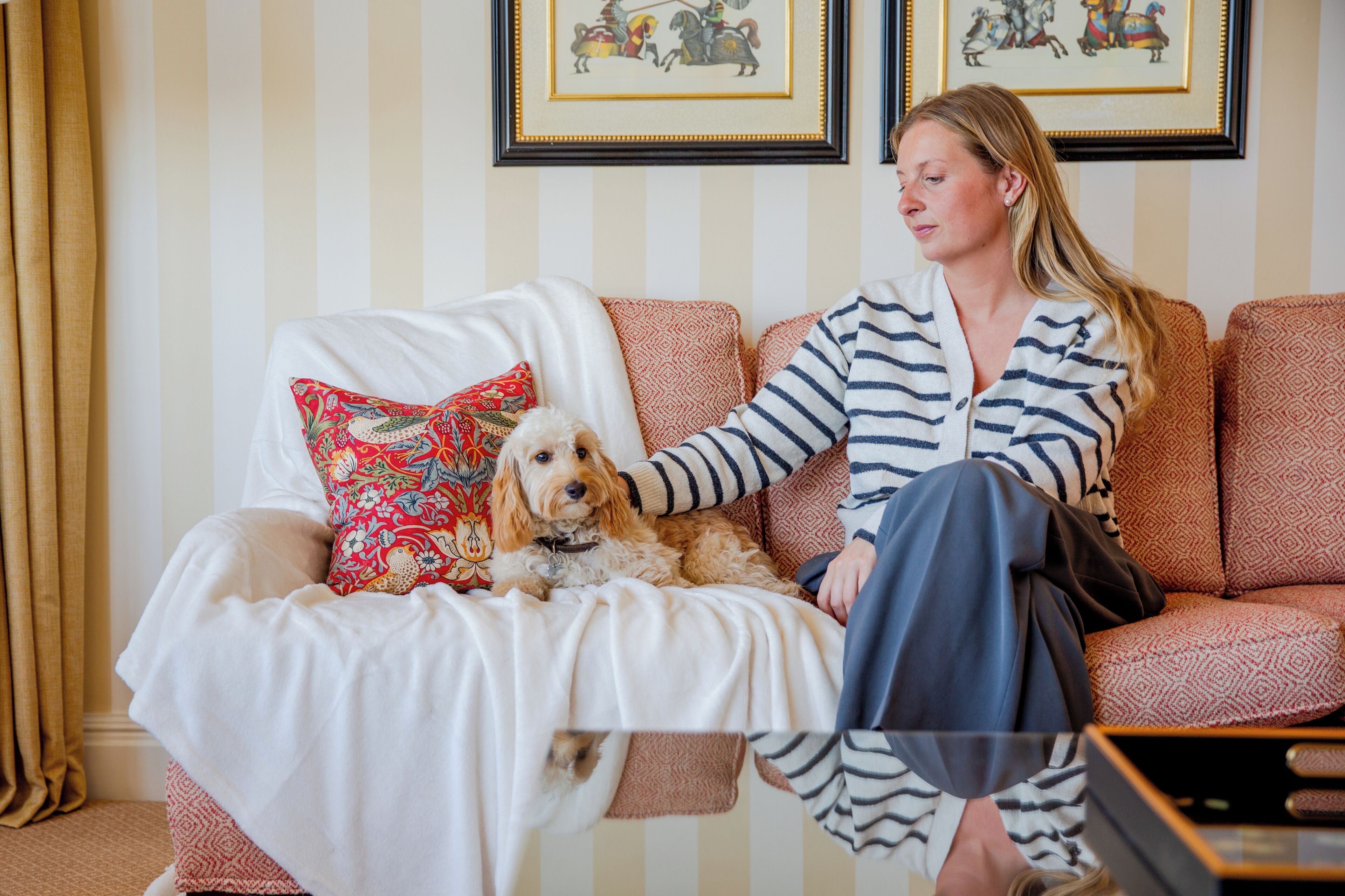 A person sitting on a patterned sofa with a dog, in a room with striped wallpaper and framed pictures on the wall.