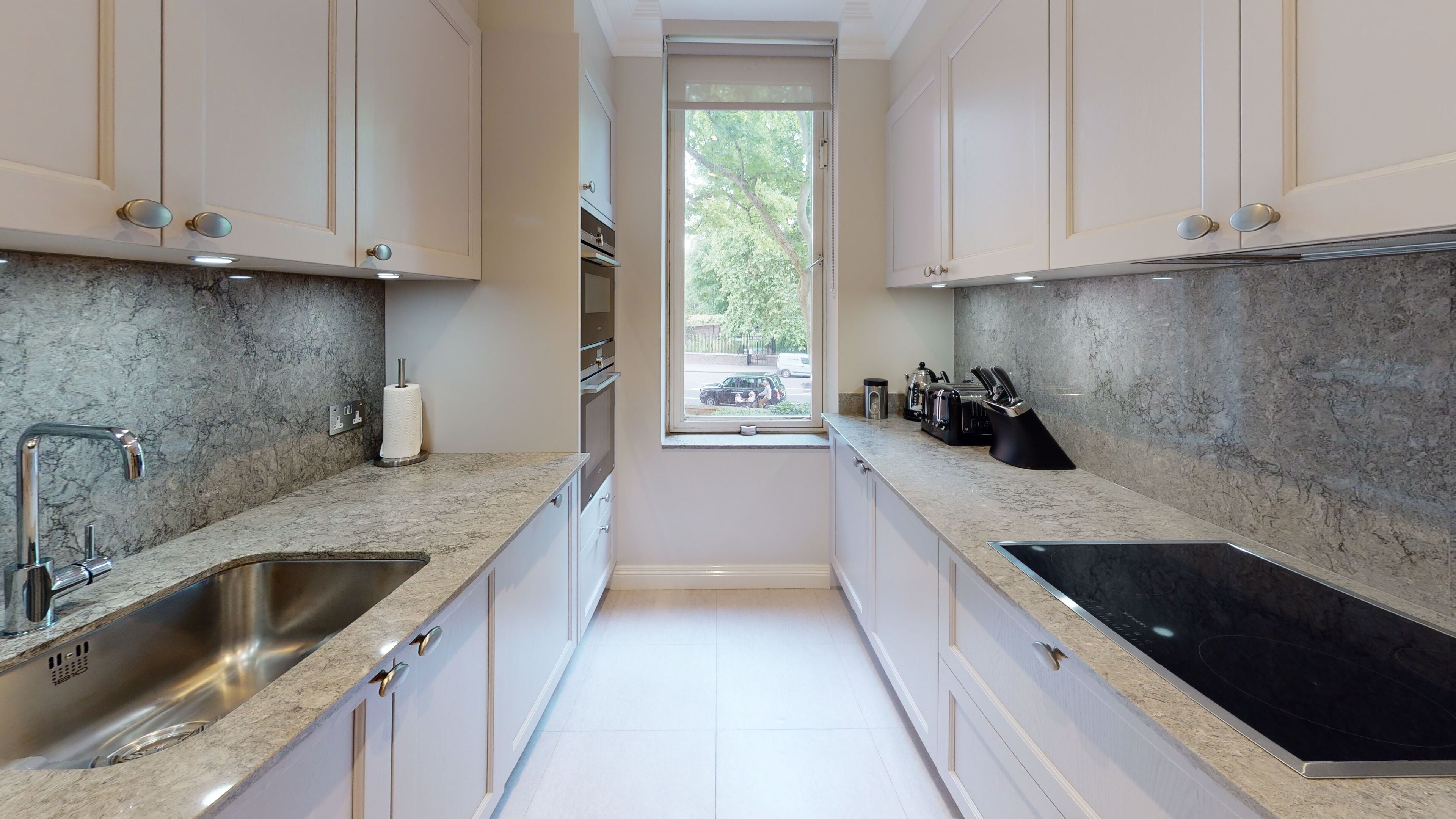 A narrow kitchen with white cabinets, granite countertops, stainless steel sinks, and a black induction cooktop.