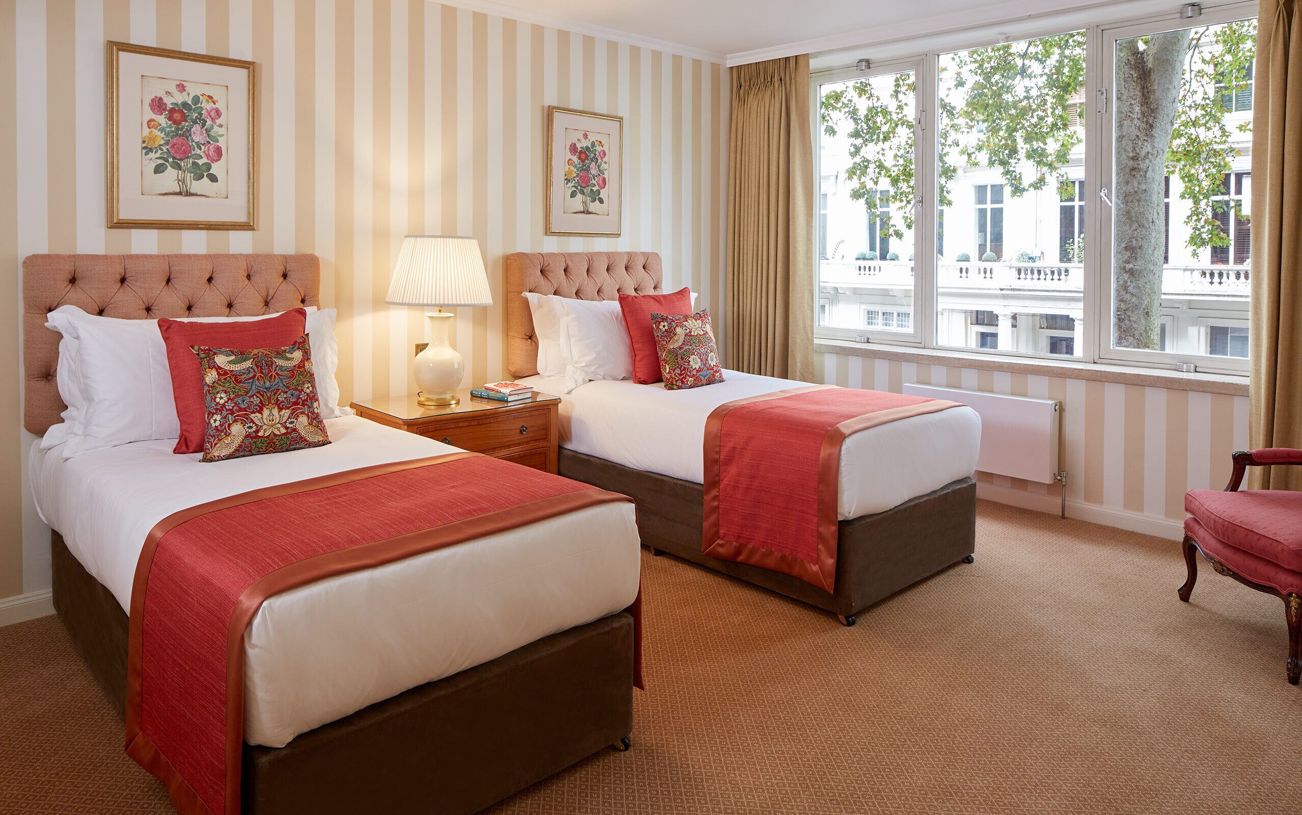 A hotel room with two beds, striped wallpaper, and a window with a view of buildings.