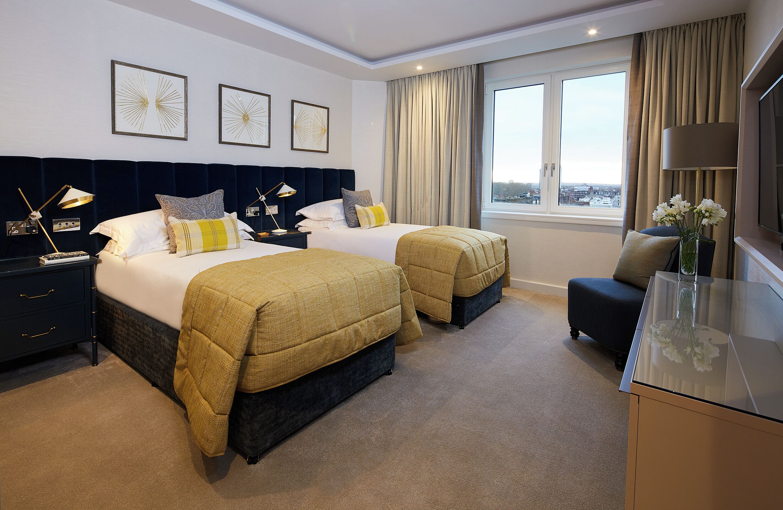 A hotel room with two beds, a desk, a chair, a TV, and a large window with curtains.