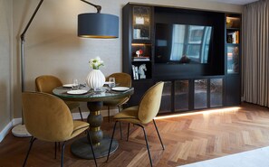 A dining area with a round glass table, four chairs, a vase with flowers, and a television on a black entertainment unit. - Cheval Gloucester Park at Kensington (London)