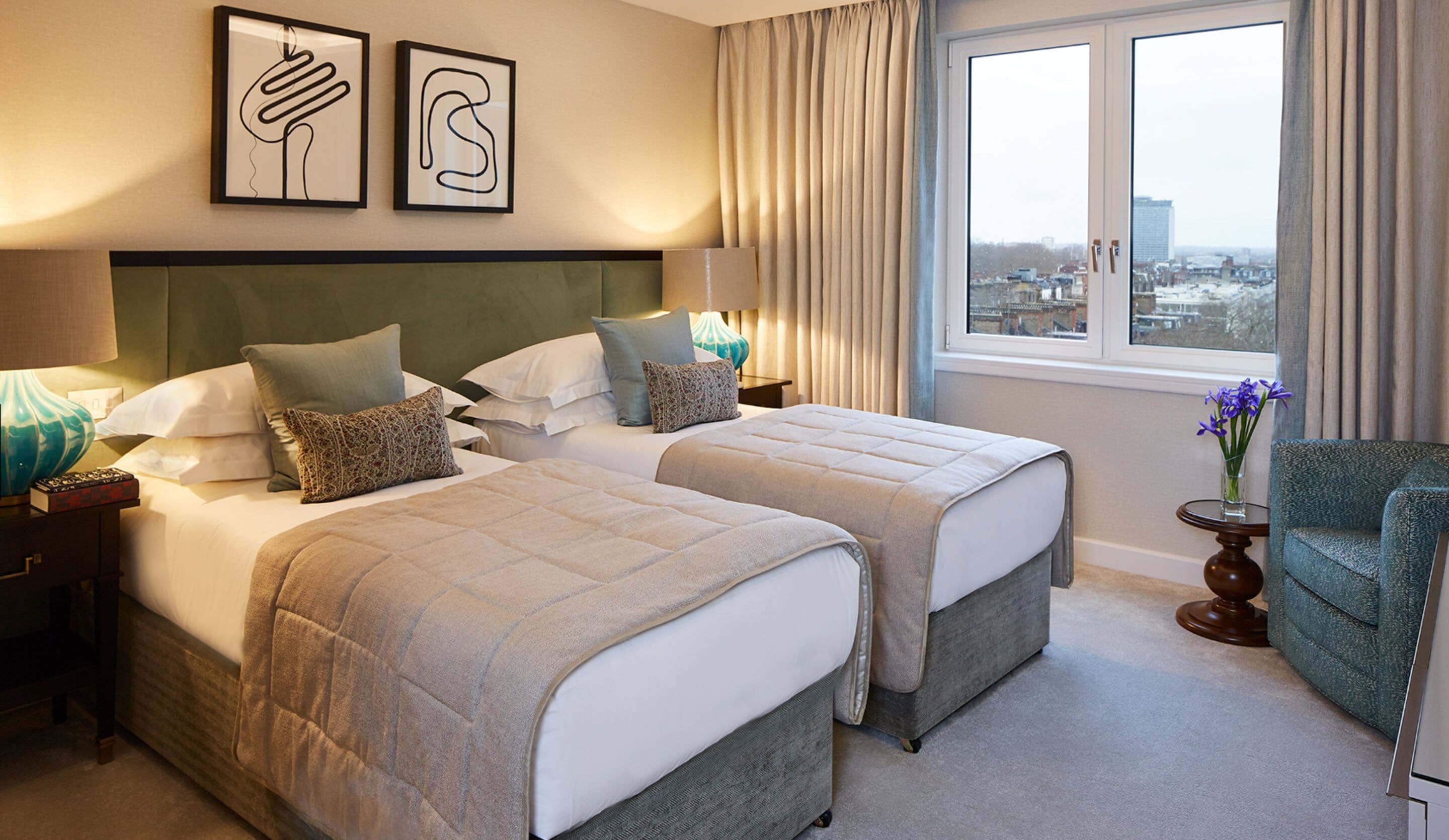 A hotel room with two beds, a view of the city through the window, and artwork on the wall.