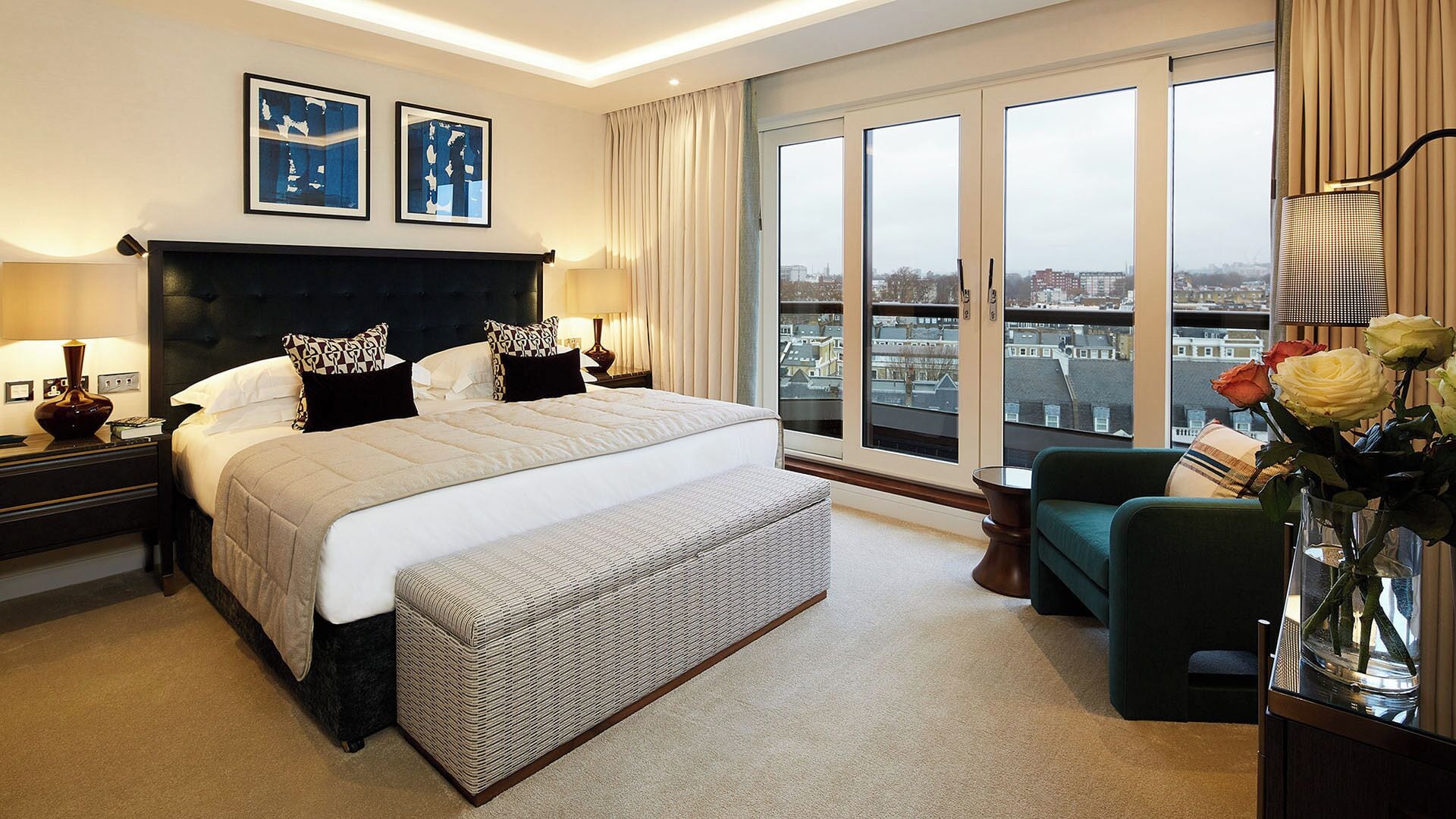 A hotel room with a large bed, two bedside tables, a chair, a vase with flowers, and a view of the city through large windows.