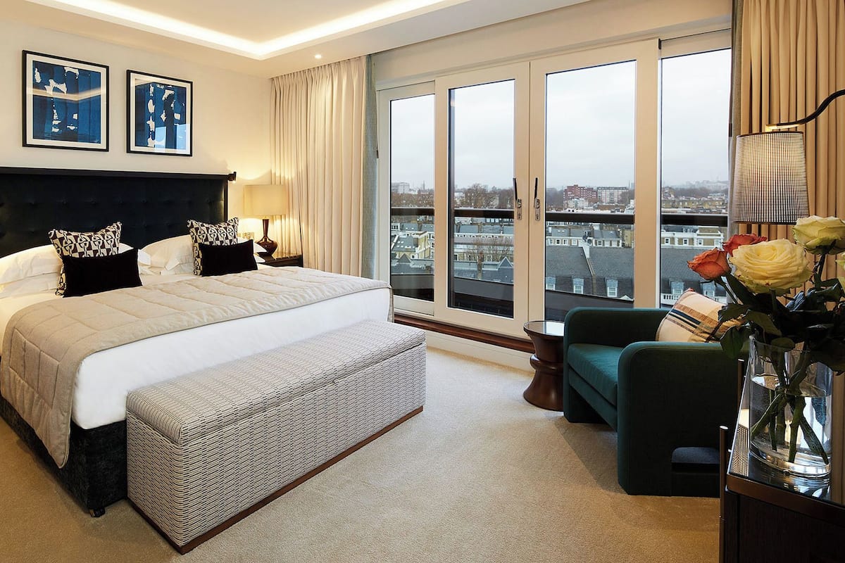Ein Hotelzimmer mit einem großen Bett, zwei Nachttischen, einem Sessel, einer Vase mit Blumen und Blick auf die Stadt durch große Fenster.