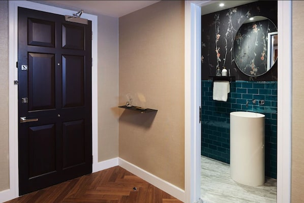 A hallway with a dark wooden door, a beige wall, and a bathroom with a blue tiled wall, a white sink, and a mirror with floral wallpaper. - Cheval Gloucester Park at Kensington (London)