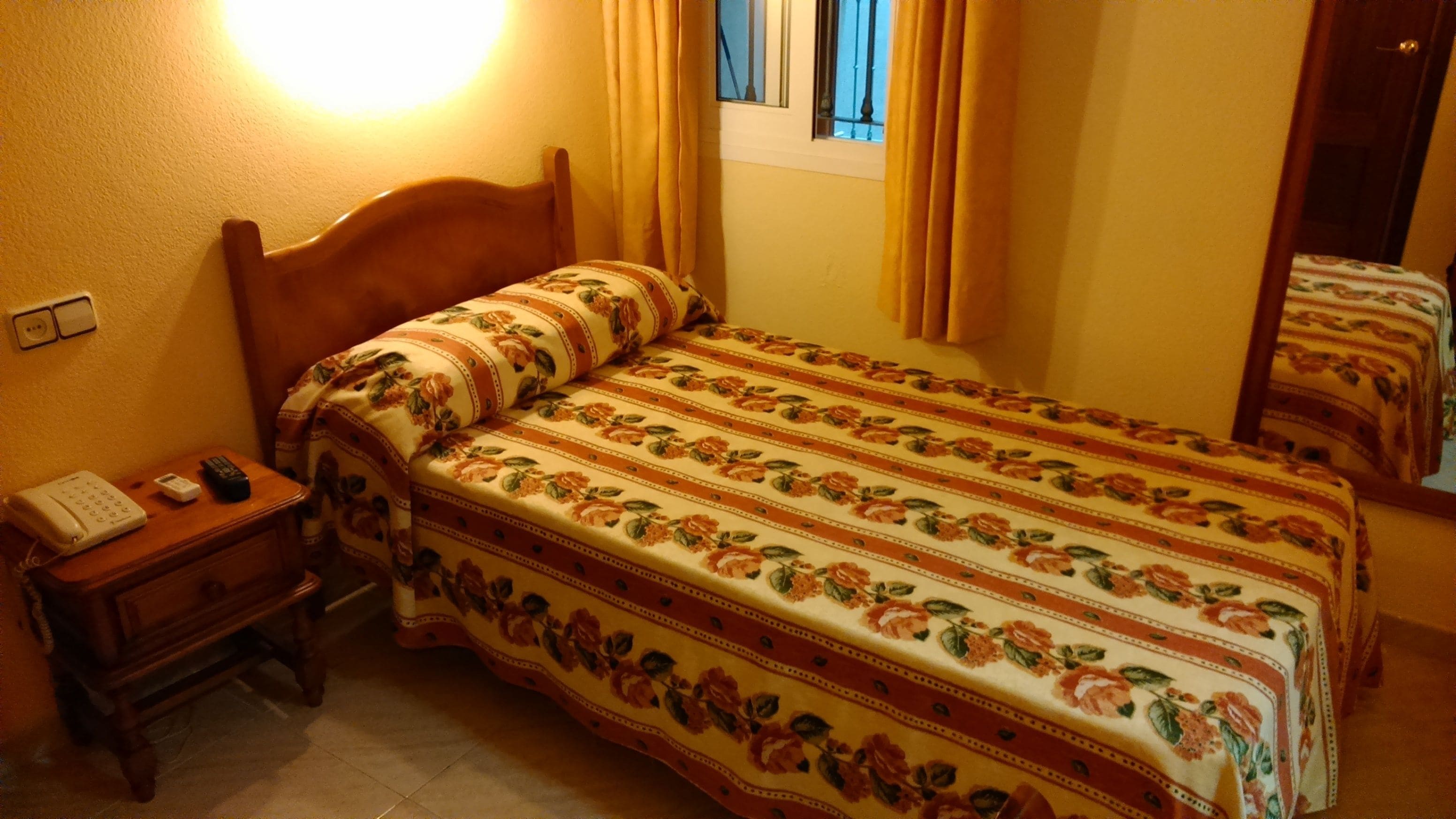 A bedroom with a wooden headboard, a bedside table with a phone and remote, and a floral-patterned bedspread.