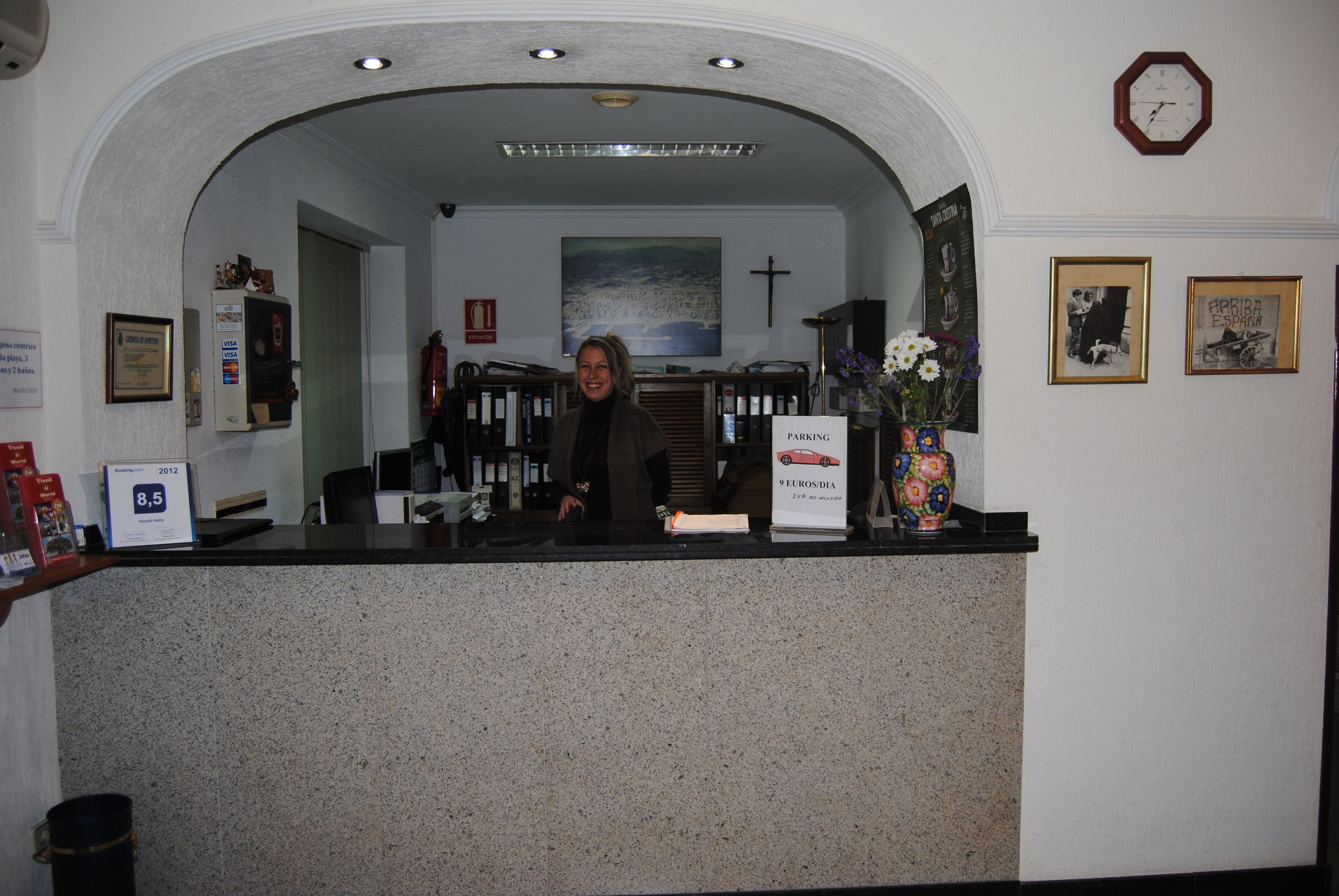 Un taulell de recepció amb un cartell de benvinguda emmarcat, un rellotge i una creu a la paret.