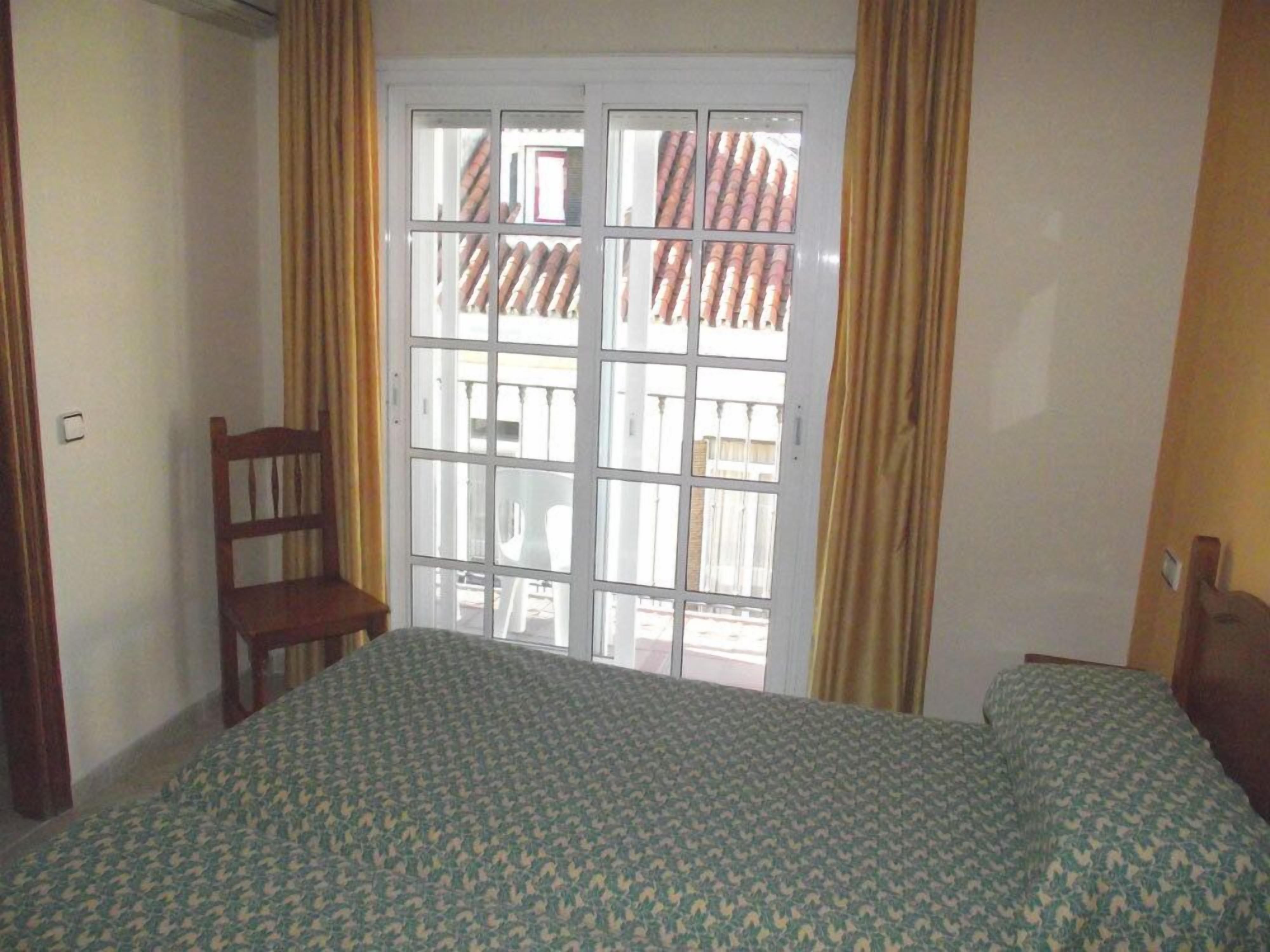 A room with a bed, a chair, and a window with a view of a building with red roofs.