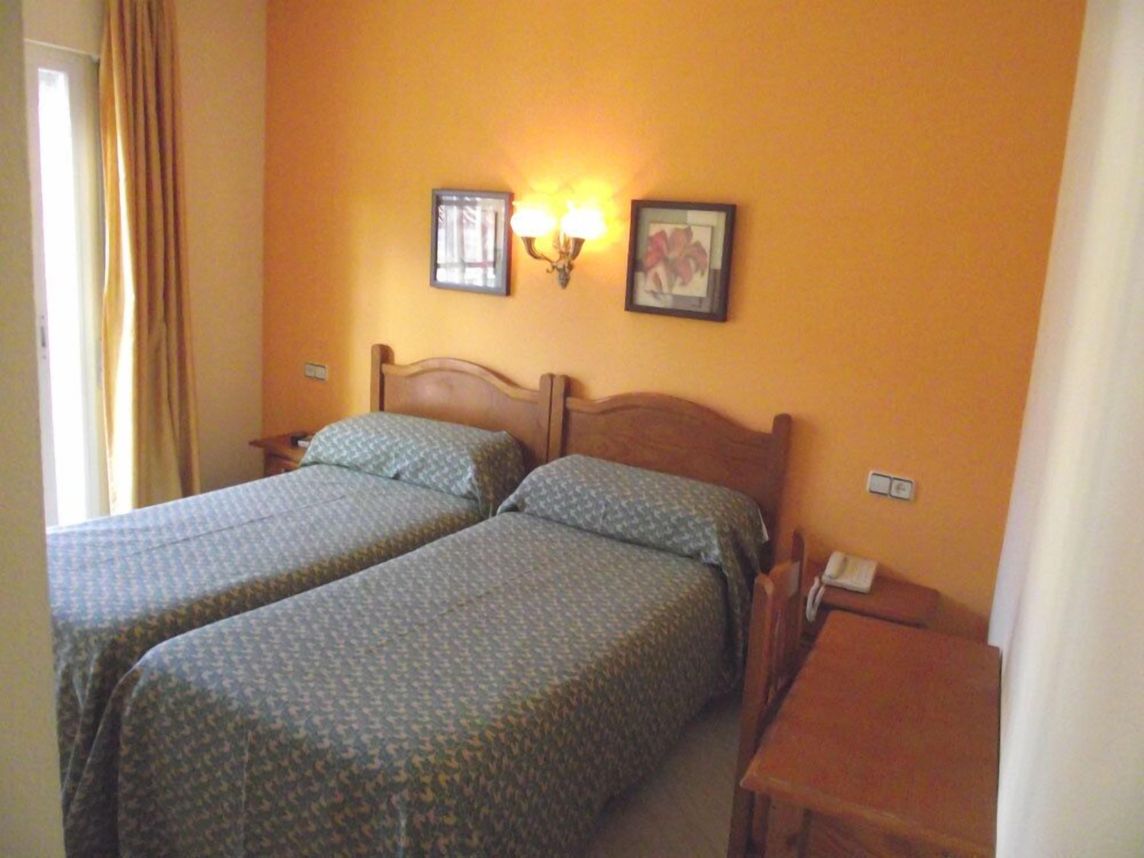 A room with two beds, a nightstand, a table, and a wall-mounted lamp.
