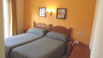 A room with two beds, a nightstand, a table, and a wall-mounted lamp.