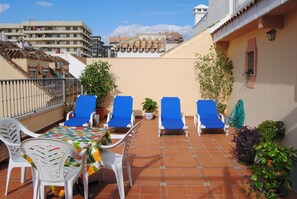 Una terraza en la azotea con cuatro sillones azules, una mesa con mantel floreado y plantas en macetas.