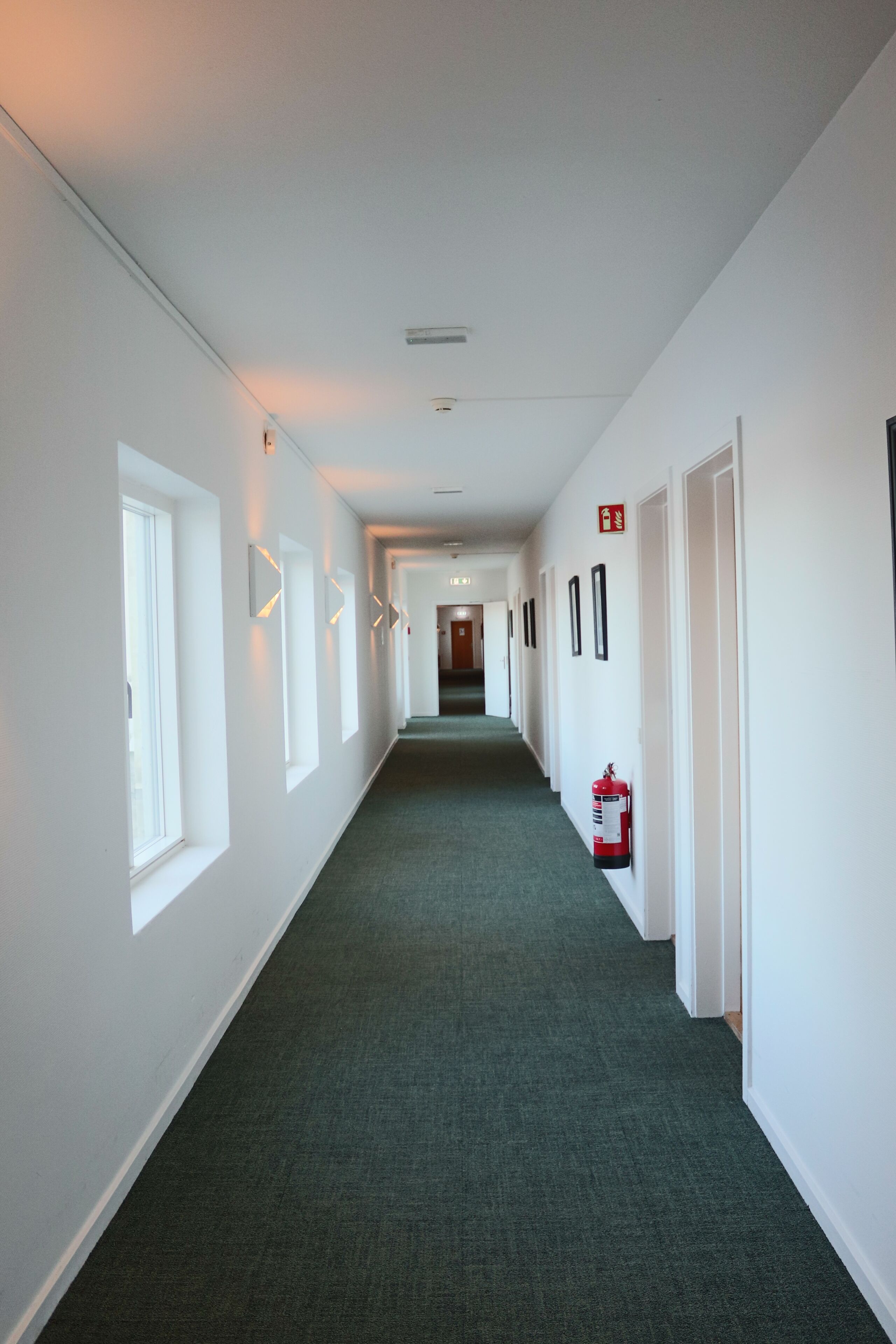A long, narrow hallway with white walls, green carpet, and doors on both sides.