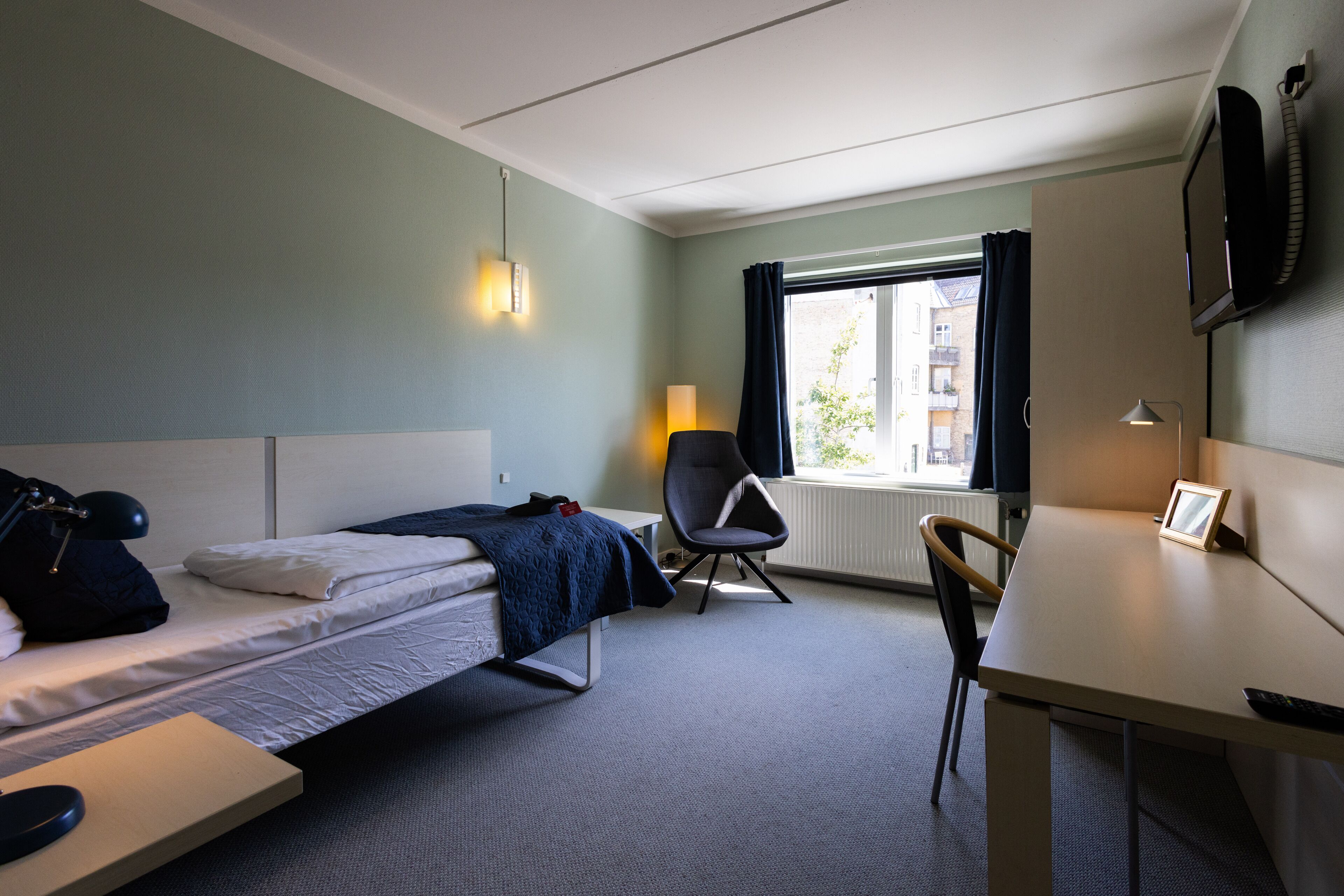 A hotel room with a bed, a desk, a chair, a lamp, and a TV.