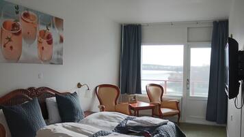 Quarto de hotel com cama, duas cadeiras, uma mesinha, televisão e uma janela grande com cortinas.