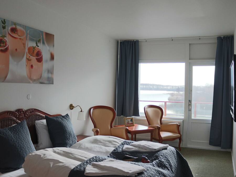 A hotel room with a bed, two chairs, a small table, a TV, and a large window with curtains.