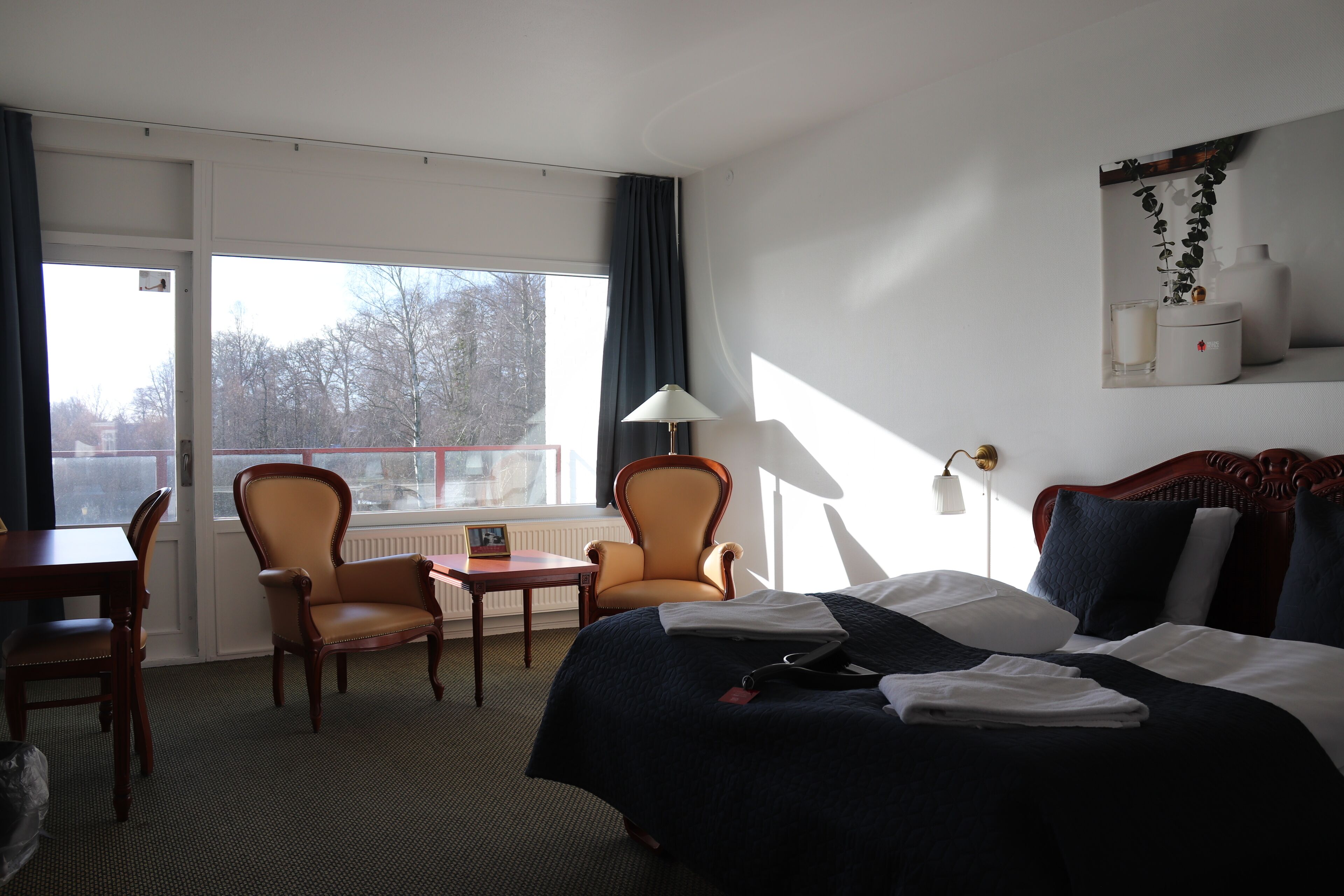 A hotel room with a large bed, two armchairs, a small table, and a view of the outdoors.