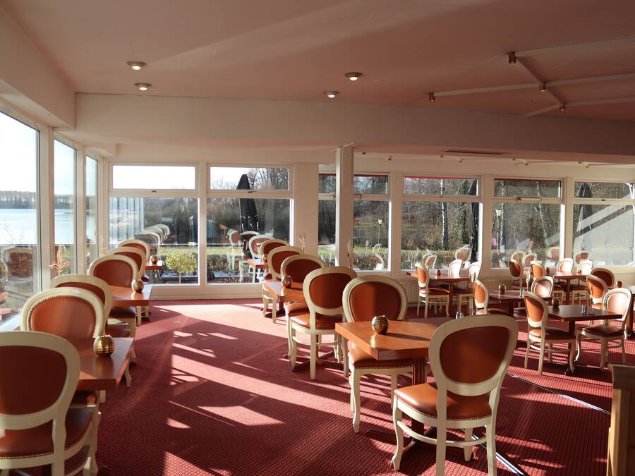 A dining area with tables and chairs arranged for guests, large windows offering views of the outdoors, and a red carpeted floor.