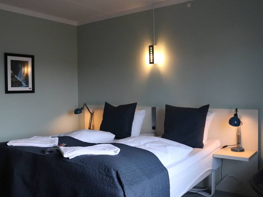 Un lit bien fait, avec des draps blancs et une couverture sombre, une lampe de chevet et un tableau encadré au mur.