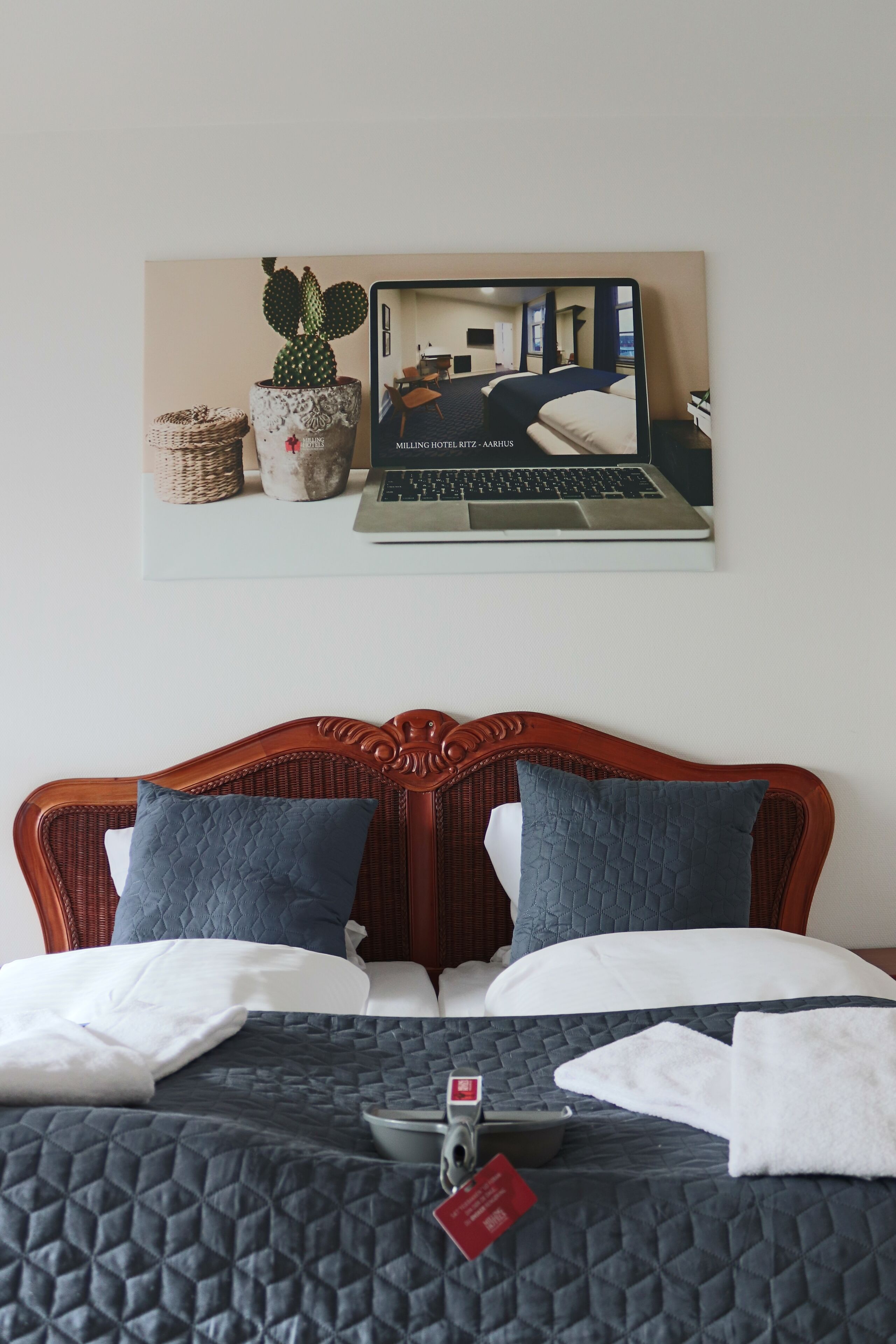 Une chambre d’hôtel avec un lit, deux oreillers, un ordinateur portable posé sur un support, une petite table et un tableau accroché au mur représentant une autre chambre.