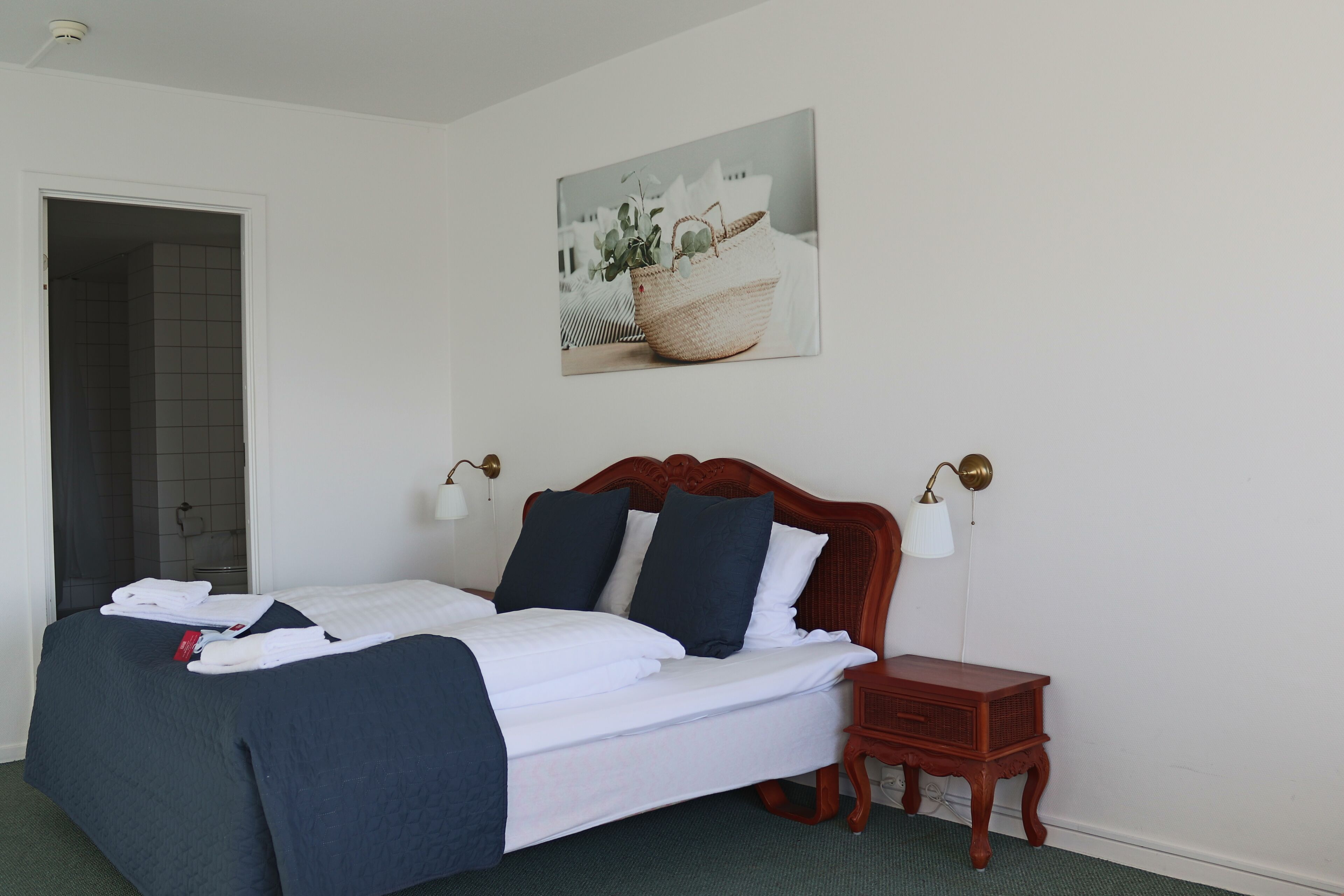 A neatly made bed with white and navy bedding, a small wooden nightstand, a wall-mounted lamp, and a framed picture above the bed.
