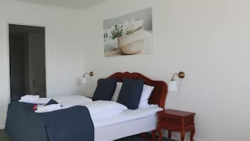 A neatly made bed with white and navy bedding, a small wooden nightstand, a wall-mounted lamp, and a framed picture above the bed.
