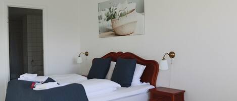A neatly made bed with white and navy bedding, a small wooden nightstand, a wall-mounted lamp, and a framed picture above the bed.