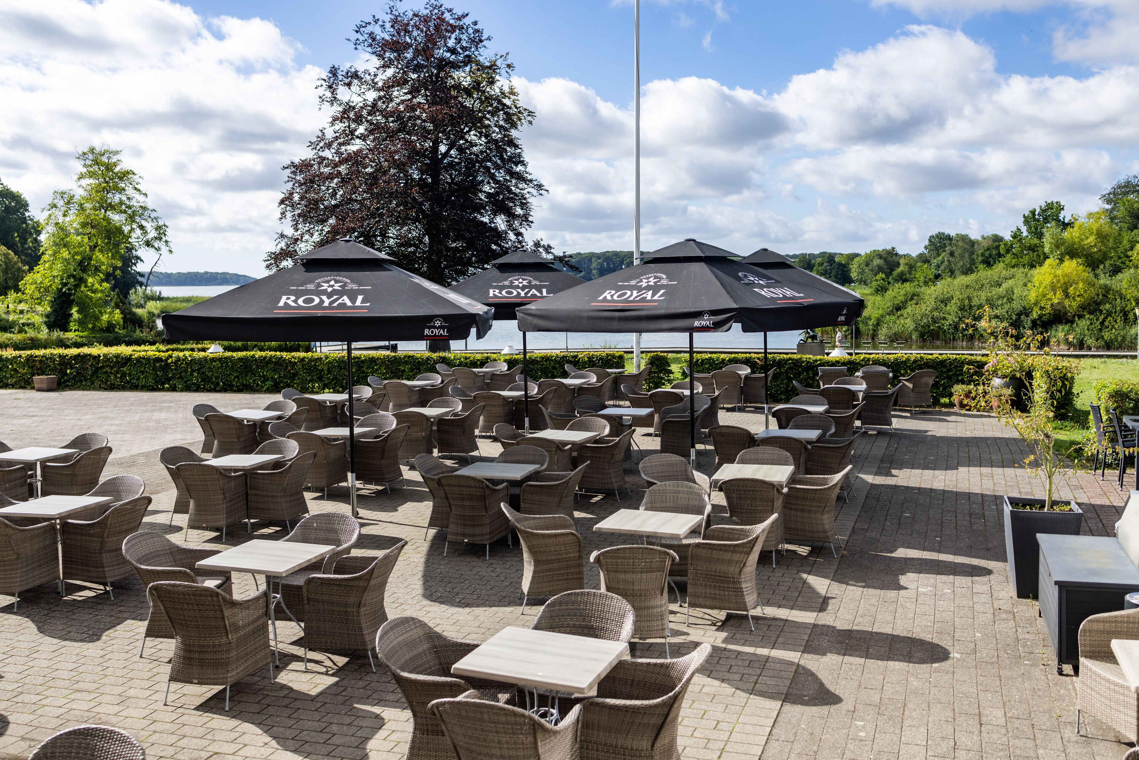 Buiten zitplaatsen met rieten stoelen en tafels, parasols en uitzicht op een meer.