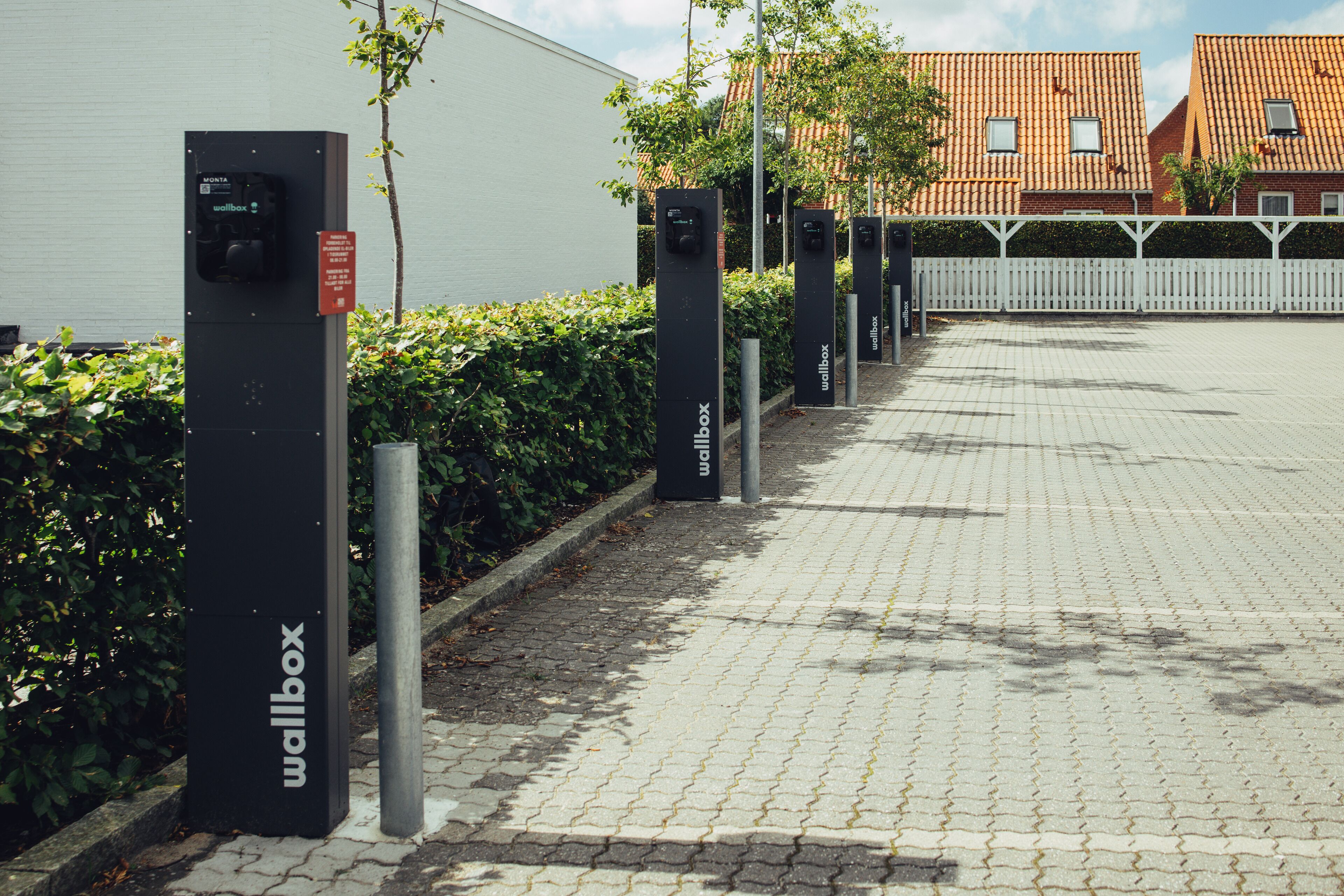 Uma fileira de estações de carregamento para carros elétricos ao longo de um caminho pavimentado, com uma cerca branca e vegetação ao fundo.