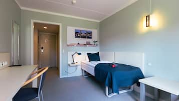 Et værelse med seng, skrivebord, stol, natbord, lampe og en vægmonteret reol med billeder.