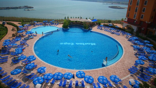 A large outdoor swimming pool with multiple lanes, surrounded by blue lounge chairs and umbrellas, with a view of a body of water and distant buildings. - Menada Ravda Apartments (Pomorie)