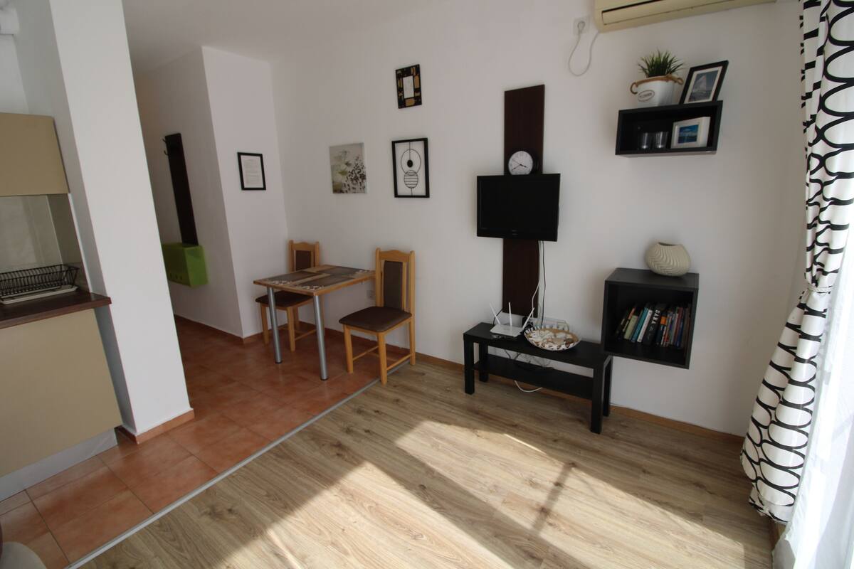 A modern living room with a wooden floor, a dining table with chairs, a TV mounted on the wall, and a bookshelf with books and decorative items.