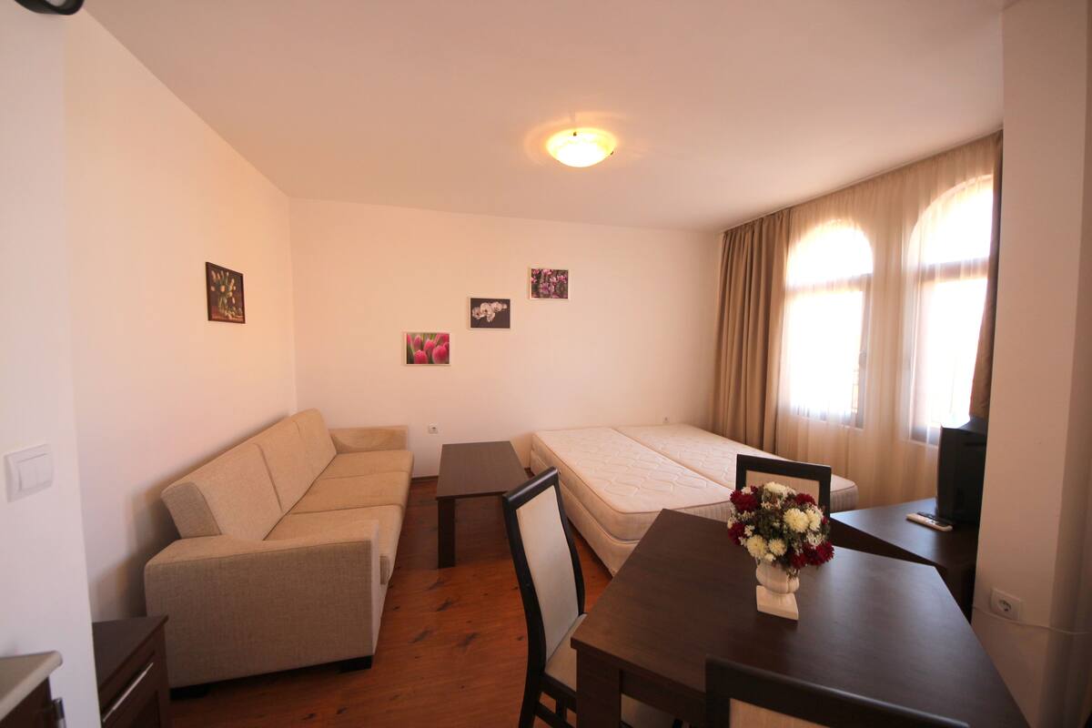 A compact room with a sofa, a single bed, a small table with a vase of flowers, and a television.