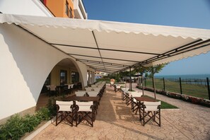 An outdoor patio with tables and chairs under a white canopy, overlooking a body of water. - Menada Ravda Apartments (Pomorie)