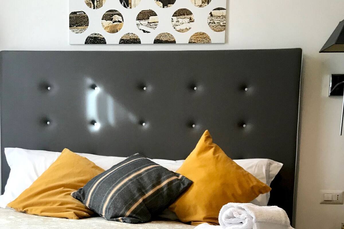 A neatly made bed with a patterned bedspread, a rolled white towel, and two pillows in mustard and striped colors.