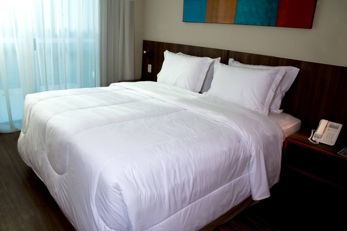 A hotel room with a large bed, a bedside telephone, and a window with sheer curtains.