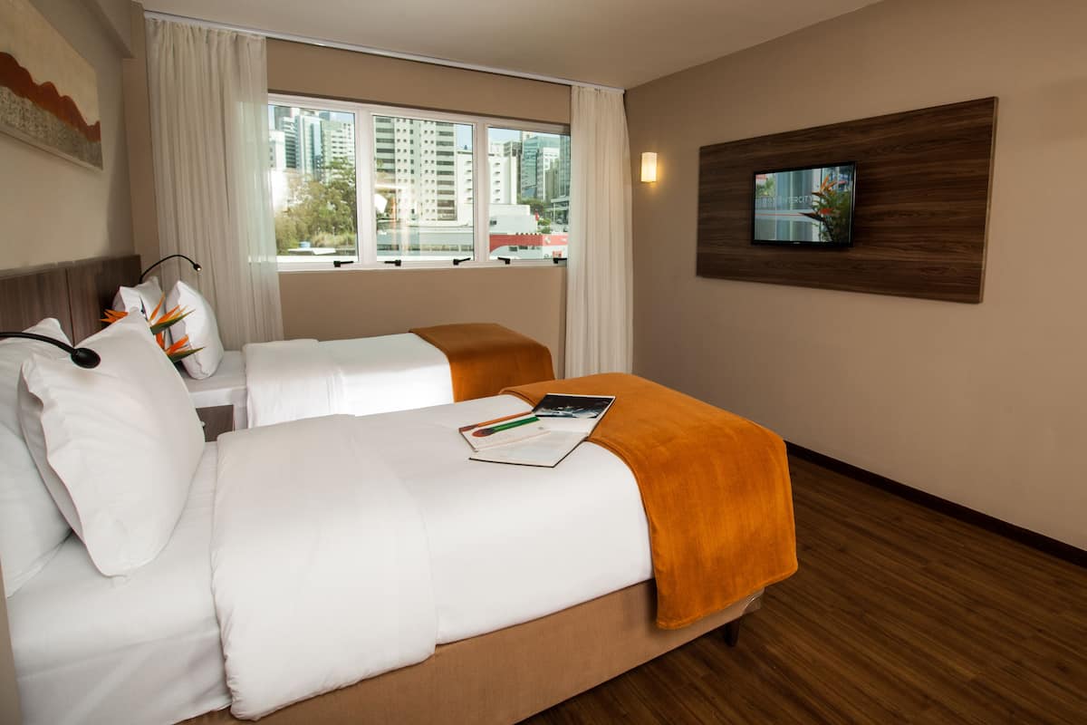 A hotel room with two beds, a TV on the wall, and a window with a city view.