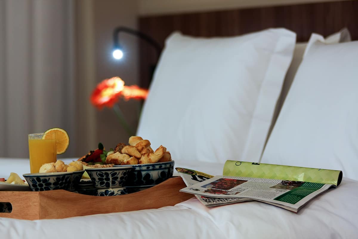 A neatly arranged breakfast tray with croissants, a glass of orange juice, and a newspaper on a bed with white linens and pillows.