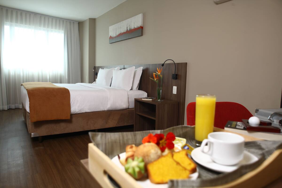 A hotel room with a bed, bedside table, and a breakfast tray with food and a cup.