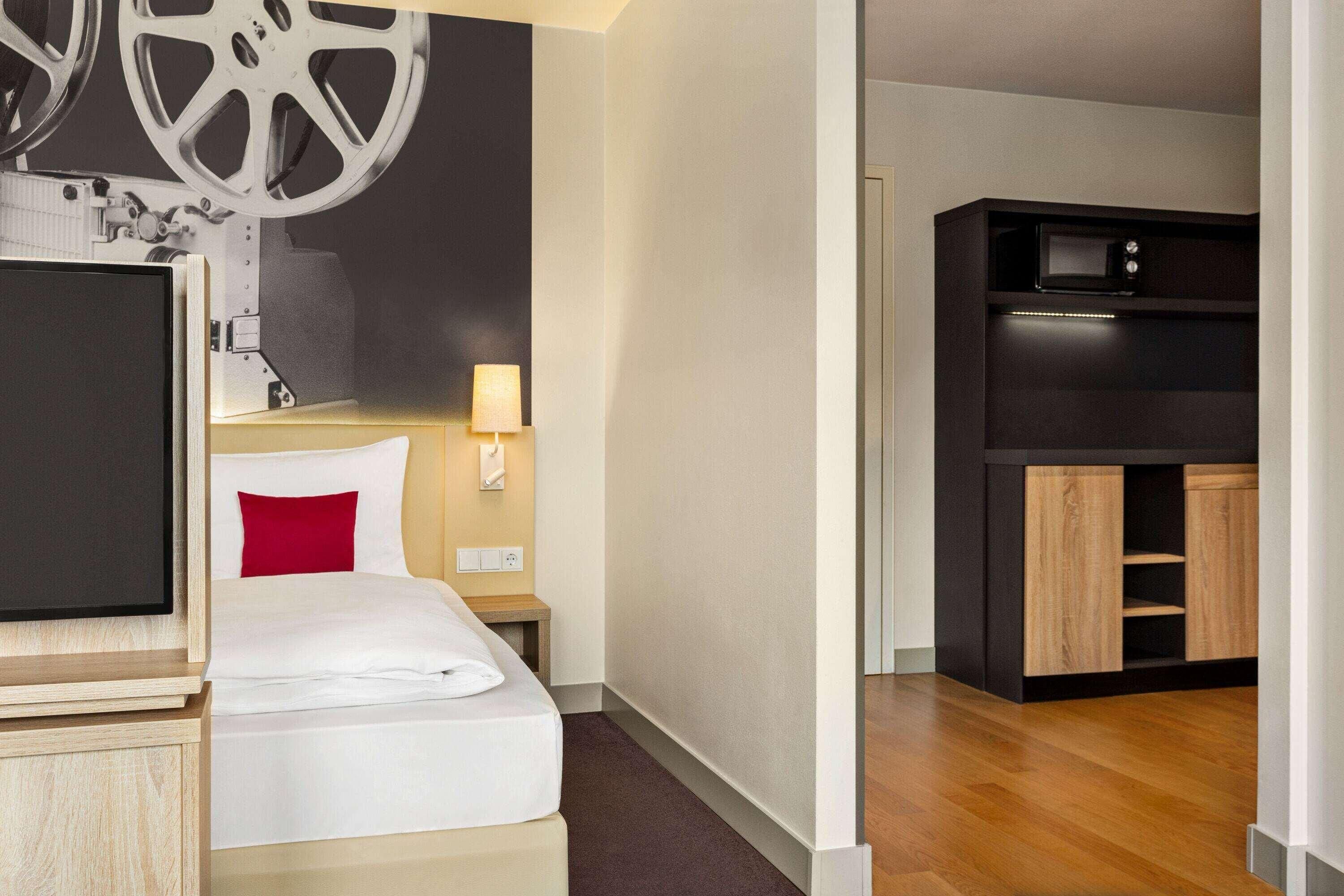 A hotel room with a bed, a TV, a desk, a lamp, and a kitchenette.