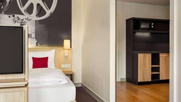 Habitación de hotel con cama, televisor, escritorio, lámpara y kitchenette.