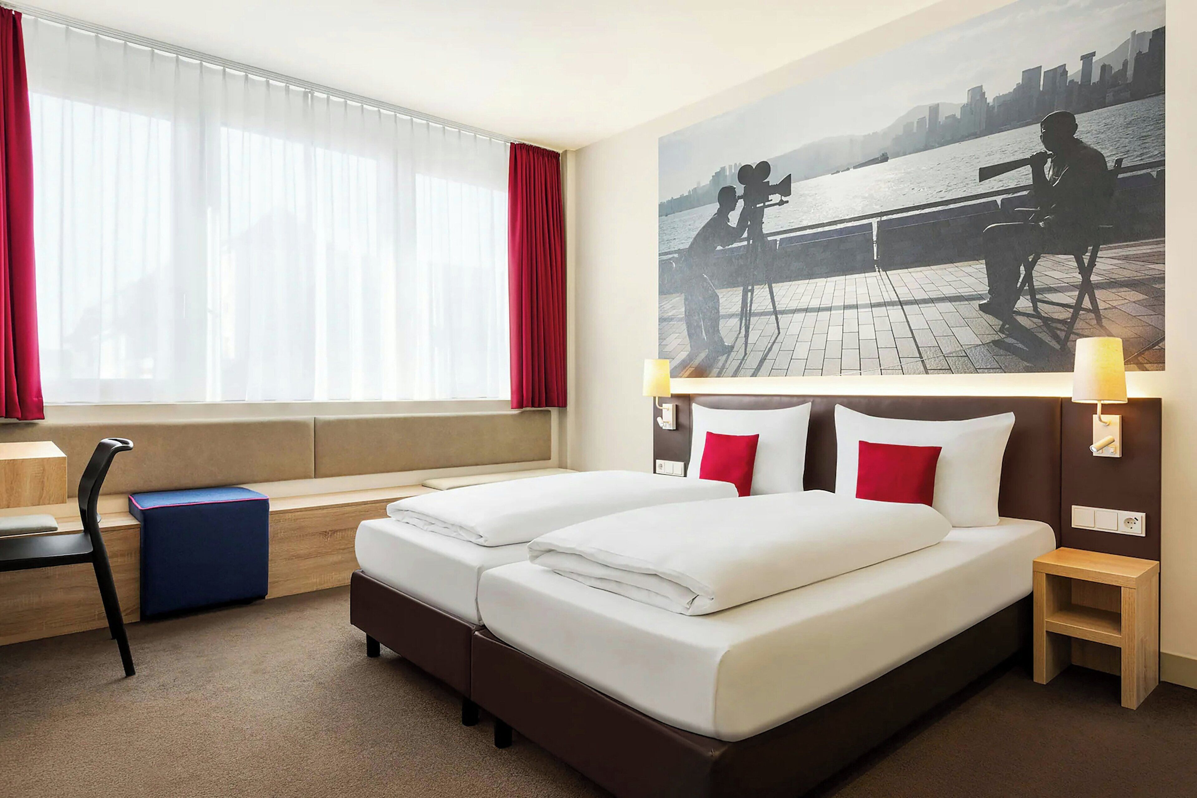 A hotel room with a large bed, a desk, a chair, a window with curtains, and a cityscape painting on the wall.