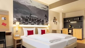A hotel room with a large bed, a desk, a chair, a small table, a microwave, and a shelf with items. - City Hotel MAX - Munich West (Munich)