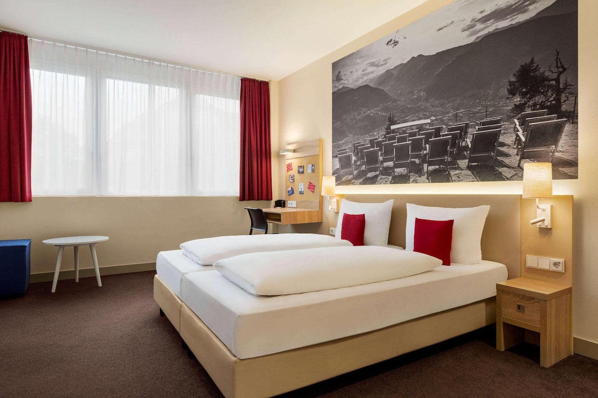 Una habitación de hotel con una cama grande, un escritorio, una silla y un ventanal con cortinas.